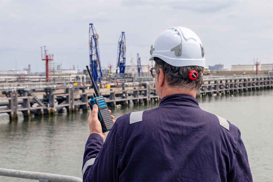 Man aan een aanlegsteiger met een portofoon in zijn linker hand. Hij draagt een donkerblauwe overal en een witte helm.